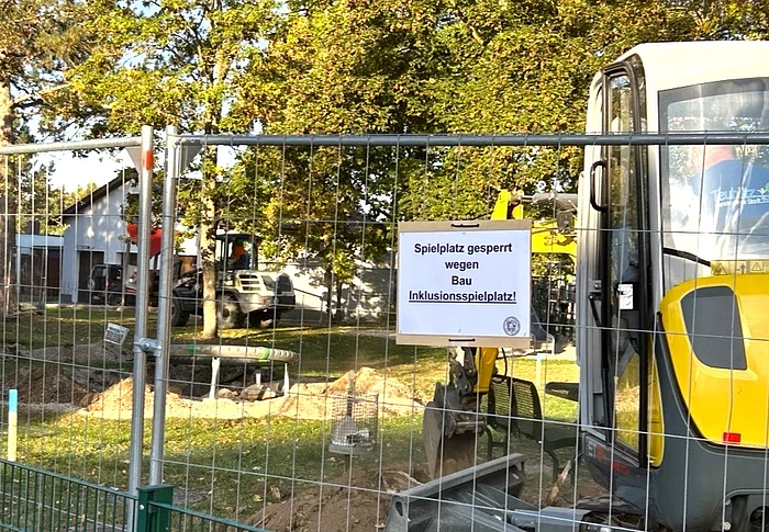 Ein abgesperrter Spielplatz mit Bauzaun, Bagger und Erdarbeiten zeigt den Beginn der Bauarbeiten für den neuen Inklusionsspielplatz.