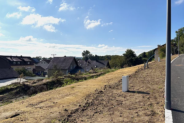 Ein leicht ansteigendes, unbebautes Grundstück mit brauner Erde liegt an einer neu asphaltierten Straße; im Hintergrund stehen einzelne Häuser am Waldrand unter blauem Himmel.