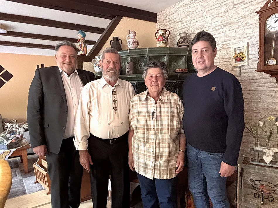 Vier Personen stehen gemeinsam in einem Wohnzimmer vor einem traditionellen grünen Kachelofen. In der Mitte steht ein älteres Ehepaar, links und rechts flankiert von zwei Männern. Einer der Männer trägt einen dunklen Anzug, die anderen legere Kleidung. Im Hintergrund sind eine Steinwand mit einer Wanduhr, Dekorationen und Holzbalken an der Decke zu sehen. Die Personen blicken in die Kamera und lächeln freundlich. Die Aufnahme vermittelt den Eindruck eines Gratulationsbesuchs anlässlich eines Ehejubiläums im familiären Umfeld.