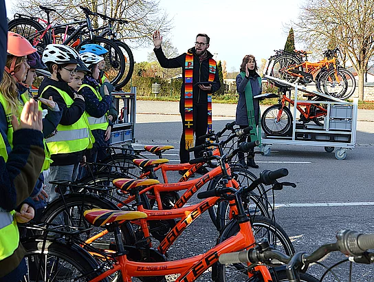 Eine Gruppe von Kindern mit Fahrradhelmen und Warnwesten steht vor ihren Fahrrädern, während ein Geistlicher die neuen Räder segnet.