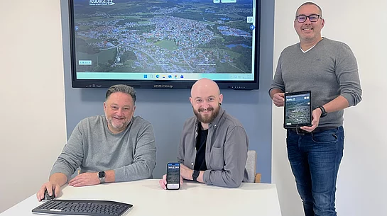 Drei Männer präsentieren im Besprechungsraum die neue Website der Stadt Teublitz. Auf einem großen Bildschirm im Hintergrund ist die Startseite der städtischen Homepage zu sehen. Einer der Männer sitzt am Tisch mit Tastatur und Maus, der zweite zeigt die Website auf seinem Smartphone, der dritte hält ein Tablet mit der geöffneten Seite. Das Bild verdeutlicht die optimierte Darstellung der neuen Website auf verschiedenen Endgeräten.