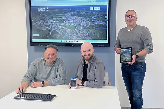 Drei Männer präsentieren im Besprechungsraum die neue Website der Stadt Teublitz. Auf einem großen Bildschirm im Hintergrund ist die Startseite der städtischen Homepage zu sehen. Einer der Männer sitzt am Tisch mit Tastatur und Maus, der zweite zeigt die Website auf seinem Smartphone, der dritte hält ein Tablet mit der geöffneten Seite. Das Bild verdeutlicht die optimierte Darstellung der neuen Website auf verschiedenen Endgeräten.