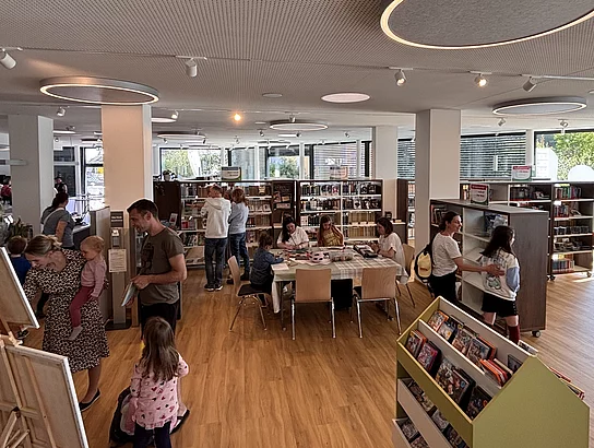 Familien und Kinder erkunden gemeinsam den offenen Innenraum der Bücherei mit Bücherregalen und Basteltisch.