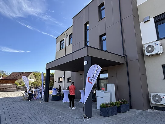 Besucherinnen und Besucher stehen vor dem Eingangsbereich des Gebäudes bei sonnigem Wetter.