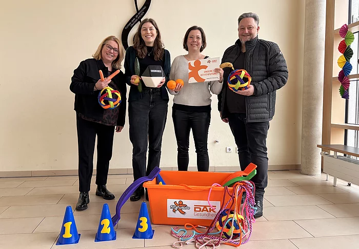 Vier Personen stehen in einem Innenraum nebeneinander, lächeln in die Kamera und präsentieren bunte Spiel- und Bewegungsmaterialien wie Bälle, Seile und Markierungshütchen aus einer großen orangefarbenen Kiste.