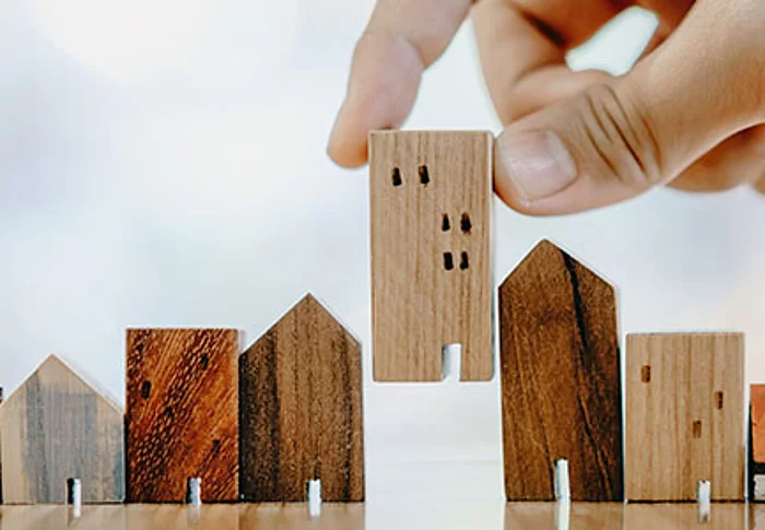 Das Foto zeigt eine Hand, die ein kleines Holzhaus zwischen weitere Holzhausmodelle stellt und damit sinnbildlich die Teilnahme von Haushalten am Mikrozensus darstellt.