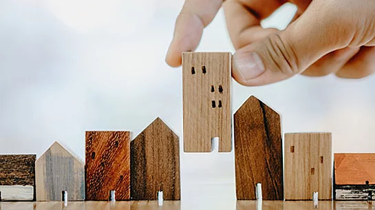 Das Foto zeigt eine Hand, die ein kleines Holzhaus zwischen weitere Holzhausmodelle stellt und damit sinnbildlich die Teilnahme von Haushalten am Mikrozensus darstellt.