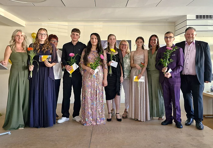 Schülerinnen und Schüler der Abschlussklasse stehen gemeinsam mit ihrer Klassenleiterin, weiteren Lehrkräften und Bürgermeister Thomas Beer auf der Bühne der Abschlussfeier. Die Absolventinnen und Absolventen halten Blumen und ihre Abschlussunterlagen in den Händen und blicken lächelnd in die Kamera.