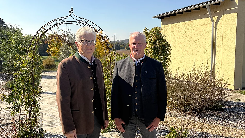 Zwei Männer stehen bei sonnigem Herbstwetter nebeneinander auf einem gepflasterten Weg in einer Gartenanlage. Links steht Robert Wutz mit weißem Haar und Brille. Er trägt eine braune Trachtenjacke über einer dunklen Weste und einem weißen Hemd. Rechts steht sein Nachfolger Georg Fleischmann mit kurzen grauen Haaren und einem kleinen grauen Kinnbart. Er trägt eine dunkelgraue Trachtenjacke mit passender Weste und grauer Hose. Hinter den beiden befindet sich ein geschwungener, mit Pflanzen bewachsener Metallbogen. Rechts ist ein hellgelbes Gebäude zu sehen. Im Hintergrund erstrecken sich Grünflächen und Bäume unter blauem Himmel.