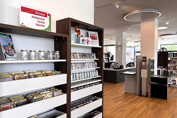 Blick in den modernen Innenraum der Stadtbücherei. Im Vordergrund befinden sich Regale mit Hörbüchern, Tonies und Medien für Kinder. Dahinter sind Selbstverbuchungsstationen, helle Säulen und offene Regalbereiche sichtbar. Runde Deckenelemente unterstreichen das moderne Design.