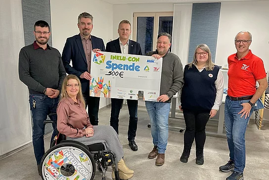 Eine Gruppe von sieben Personen präsentiert gemeinsam einen großen Spendenscheck über 500 Euro, wobei eine Frau im Rollstuhl vorne sitzt und alle freundlich in die Kamera lächeln.