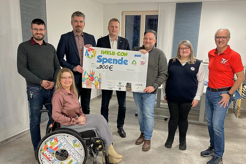 Eine Gruppe von sieben Personen präsentiert gemeinsam einen großen Spendenscheck über 500 Euro, wobei eine Frau im Rollstuhl vorne sitzt und alle freundlich in die Kamera lächeln.