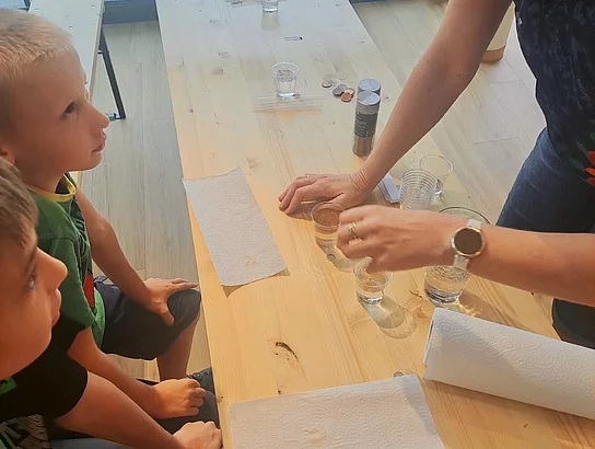 Eine Frau führt zwei Kindern an einem Holztisch ein Experiment mit kleinen Gläsern, Wasser und weiteren Materialien vor. Die Kinder beobachten aufmerksam.