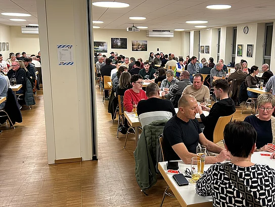 In einem großen, hell beleuchteten Saal sitzen viele Menschen dicht an dicht an Tischen, spielen Karten und unterhalten sich, während Getränke auf den Tischen stehen.