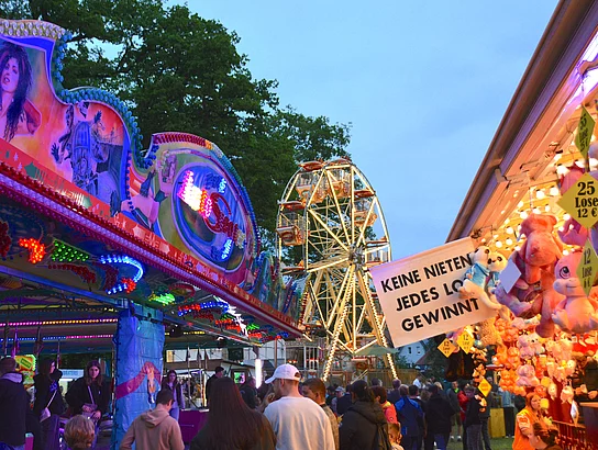 Eine zulaufende Gasse am Volksfestplatz mit bunt leuchtenden Verkaufsfenstern und einem erstrahltem Riesenrad im Hintergrund.