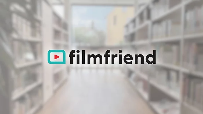 Unscharfer Bibliothekshintergrund mit Bücherregalen und großem Fenster. Im Zentrum steht das Logo „filmfriend“ in schwarzer Schrift. Links daneben befindet sich ein türkisfarbener Rahmen mit einem roten Play-Symbol – ein Hinweis auf ein digitales Film-Streaming-Angebot der Stadtbücherei.