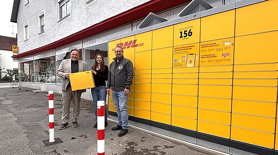 Drei Personen stehen vor der neuen gelben DHL-Packstation Nummer 156 an der Postfiliale beziehungsweise AVIA-Tankstelle in der Regensburger Straße in Teublitz. Eine Person hält symbolisch ein großes DHL-Paket in den Händen. Im Hintergrund ist die lange Paketstation mit zahlreichen Fächern zu sehen.