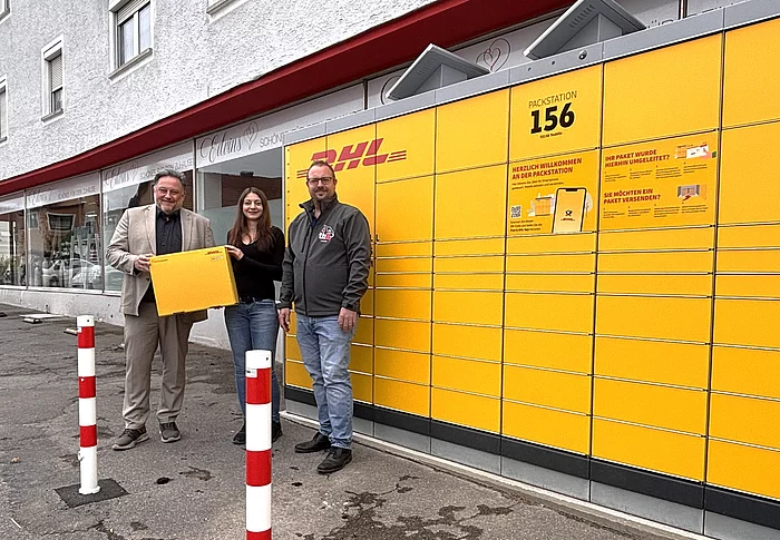 Drei Personen stehen vor der neuen gelben DHL-Packstation Nummer 156 an der Postfiliale beziehungsweise AVIA-Tankstelle in der Regensburger Straße in Teublitz. Eine Person hält symbolisch ein großes DHL-Paket in den Händen. Im Hintergrund ist die lange Paketstation mit zahlreichen Fächern zu sehen.