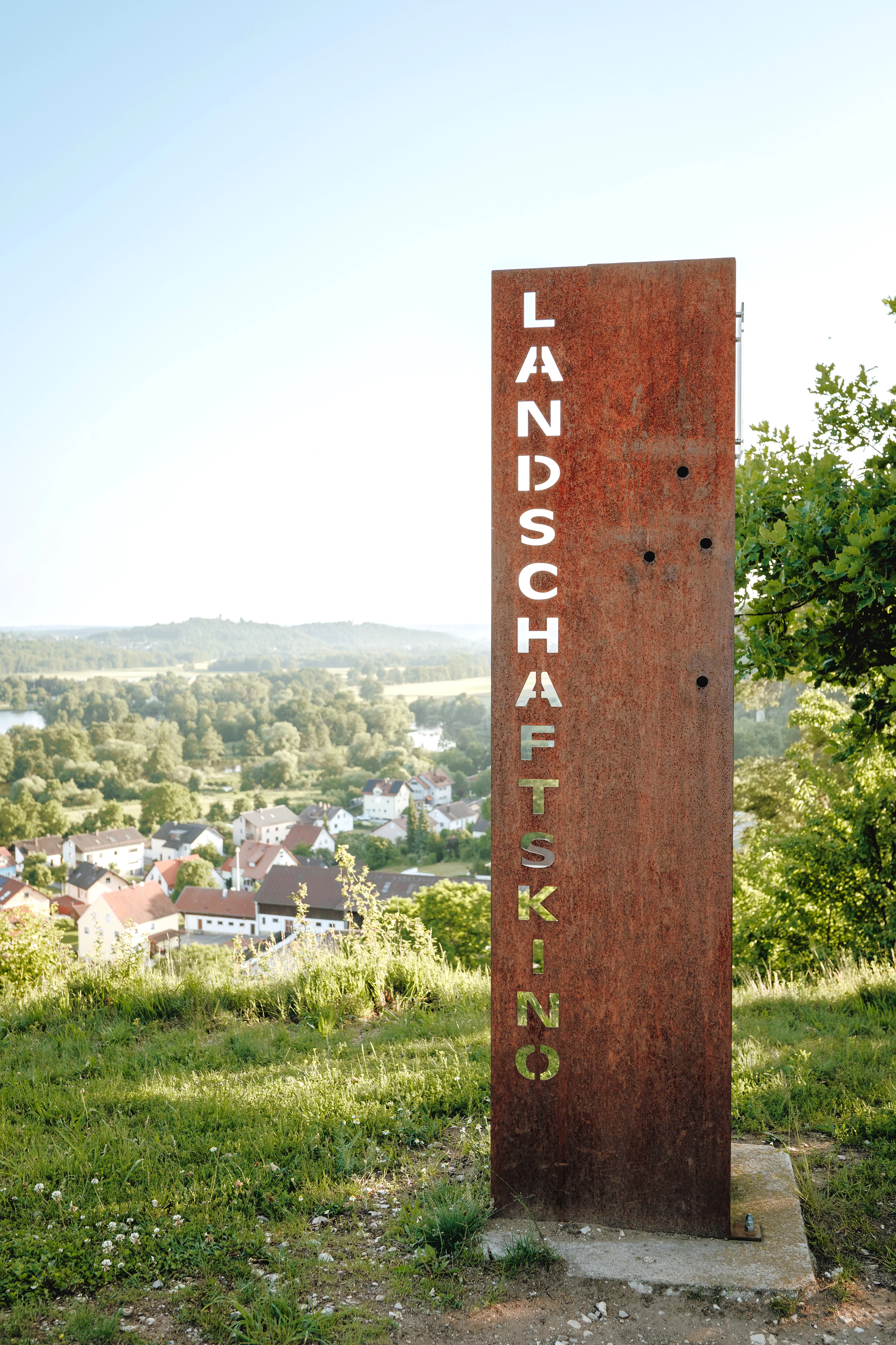 Eine Eisenstehle in Rostoptik. Vertikal ausgestanzt ist das Wort "Landschasftskino". Im Hintergrund das Ortspanorama von Premberg.