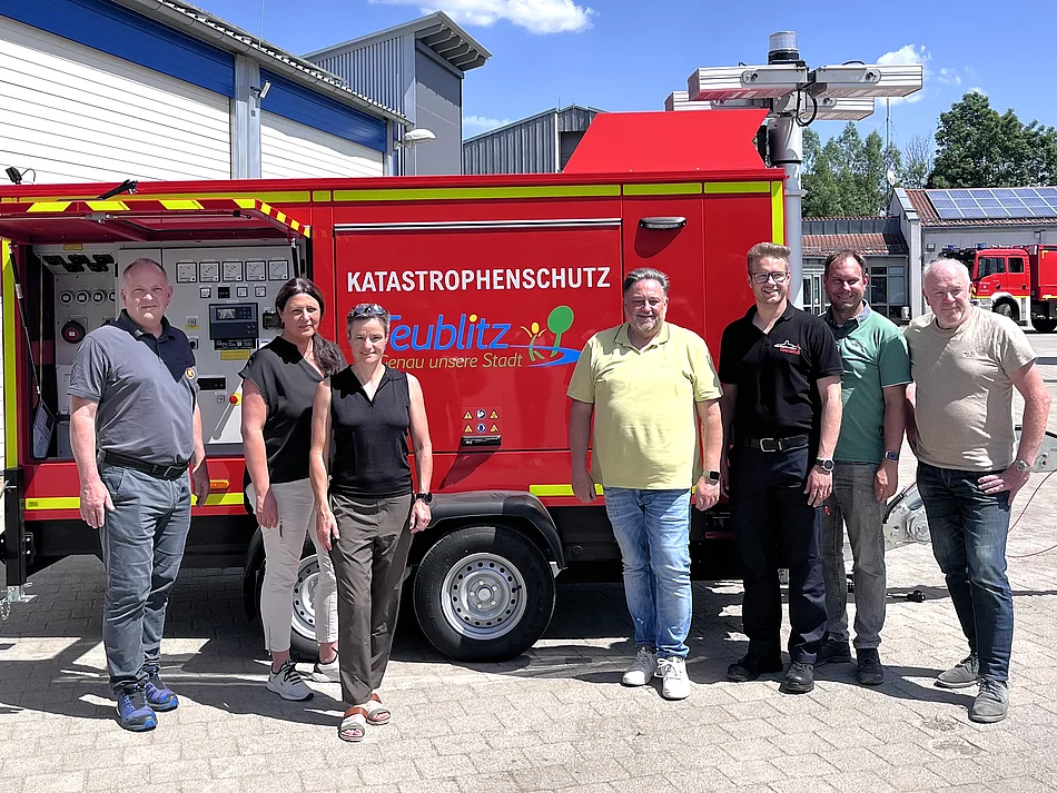 Sieben Personen stehen bei sonnigem Wetter vor einem roten mobilen Notstromaggregat mit der Aufschrift „Katastrophenschutz Teublitz“. Das Aggregat befindet sich auf einem Anhänger vor einem Gebäude des Wasserwerks beziehungsweise Feuerwehrgeländes. Im Hintergrund sind weitere Gebäude, ein Feuerwehrfahrzeug und ein blauer Himmel mit großen Wolken zu sehen.