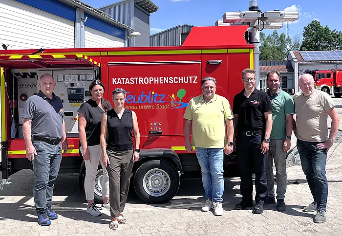 Sieben Personen stehen bei sonnigem Wetter vor einem roten mobilen Notstromaggregat mit der Aufschrift „Katastrophenschutz Teublitz“. Das Aggregat befindet sich auf einem Anhänger vor einem Gebäude des Wasserwerks beziehungsweise Feuerwehrgeländes. Im Hintergrund sind weitere Gebäude, ein Feuerwehrfahrzeug und ein blauer Himmel mit großen Wolken zu sehen.