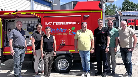 Sieben Personen stehen bei sonnigem Wetter vor einem roten mobilen Notstromaggregat mit der Aufschrift „Katastrophenschutz Teublitz“. Das Aggregat befindet sich auf einem Anhänger vor einem Gebäude des Wasserwerks beziehungsweise Feuerwehrgeländes. Im Hintergrund sind weitere Gebäude, ein Feuerwehrfahrzeug und ein blauer Himmel mit großen Wolken zu sehen.