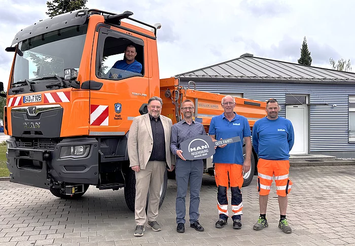Mehrere Personen stehen vor einem neuen orangefarbenen MAN-Kommunalfahrzeug des städtischen Bauhofs. Ein Mitarbeiter sitzt im Führerhaus, während Vertreter der Stadt, der Herstellerfirma und Bauhofmitarbeiter vor dem Fahrzeug für ein Übergabefoto posieren. Der neue LKW ist auf dem Gelände des Bauhofs abgestellt.