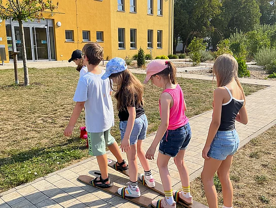 Fünf Kinder balancieren gemeinsam auf langen Team-Skiern über den Weg vor dem Mehrgenerationenhaus.
