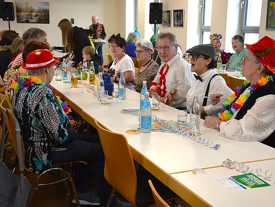 An einem langen Tisch unterhalten sich ältere Menschen lebhaft, einige singen oder sprechen laut und tragen bunte Girlanden und Hüte.