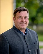 Ein Mann mit kurzem, seitlich gekämmtem Haar trägt eine traditionelle, hochgeschlossene Jacke und lächelt freundlich in die Kamera.