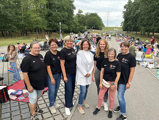 Sieben Frauen stehen lächelnd für ein Gruppenfoto auf der Straße. Sechs von ihnen tragen schwarze T-Shirts des Eltern-Kind-Treffs Maxhütte-Haidhof. Im Hintergrund erstreckt sich der gut besuchte Kinderwarenflohmarkt mit zahlreichen Verkaufsständen und Familien entlang der Straße.