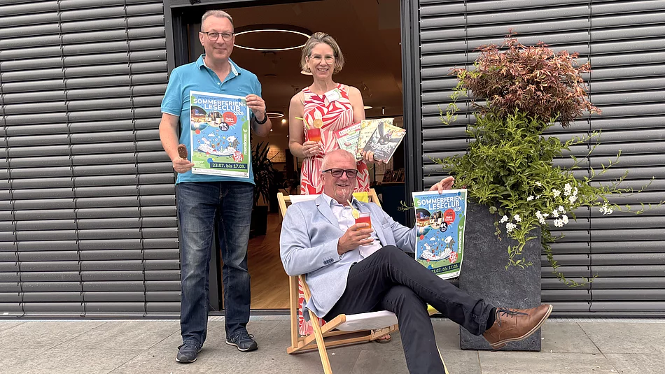 Drei Personen präsentieren vor dem Eingang der Stadtbücherei den Sommerferien-Leseclub 2026. Eine Person sitzt entspannt in einem Liegestuhl und hält ein Plakat der Aktion sowie ein sommerliches Getränk in der Hand. Die beiden weiteren Personen stehen dahinter, zeigen ebenfalls das Veranstaltungsplakat und präsentieren mehrere Kinder- und Jugendbücher sowie ein Getränk. Rechts neben der Gruppe steht ein großer Pflanzkübel mit einem Zierahorn und weißen Blüten. Die fröhliche Szene vermittelt Sommerstimmung und lädt zum Mitmachen ein.