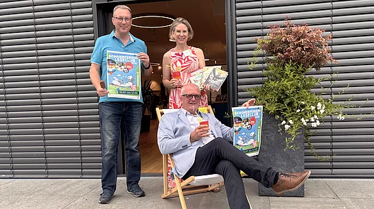 Drei Personen präsentieren vor dem Eingang der Stadtbücherei den Sommerferien-Leseclub 2026. Eine Person sitzt entspannt in einem Liegestuhl und hält ein Plakat der Aktion sowie ein sommerliches Getränk in der Hand. Die beiden weiteren Personen stehen dahinter, zeigen ebenfalls das Veranstaltungsplakat und präsentieren mehrere Kinder- und Jugendbücher sowie ein Getränk. Rechts neben der Gruppe steht ein großer Pflanzkübel mit einem Zierahorn und weißen Blüten. Die fröhliche Szene vermittelt Sommerstimmung und lädt zum Mitmachen ein.
