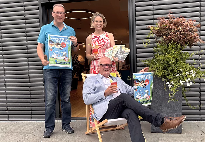 Drei Personen präsentieren vor dem Eingang der Stadtbücherei den Sommerferien-Leseclub 2026. Eine Person sitzt entspannt in einem Liegestuhl und hält ein Plakat der Aktion sowie ein sommerliches Getränk in der Hand. Die beiden weiteren Personen stehen dahinter, zeigen ebenfalls das Veranstaltungsplakat und präsentieren mehrere Kinder- und Jugendbücher sowie ein Getränk. Rechts neben der Gruppe steht ein großer Pflanzkübel mit einem Zierahorn und weißen Blüten. Die fröhliche Szene vermittelt Sommerstimmung und lädt zum Mitmachen ein.