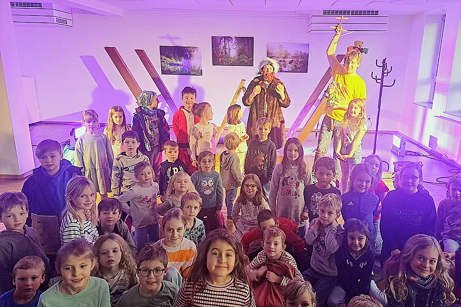 Eine große Gruppe fröhlicher Kinder sitzt und steht in einem bunt beleuchteten Raum, während zwei verkleidete Personen auf einer kleinen Bühne im Hintergrund ein Piratenstück aufführen.