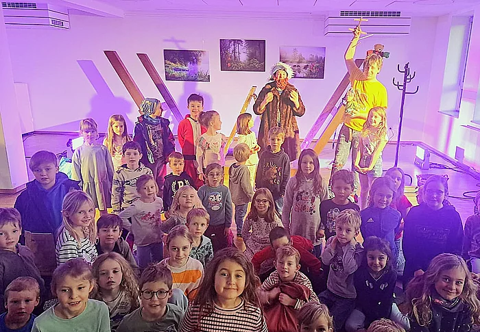 Eine große Gruppe fröhlicher Kinder sitzt und steht in einem bunt beleuchteten Raum, während zwei verkleidete Personen auf einer kleinen Bühne im Hintergrund ein Piratenstück aufführen.