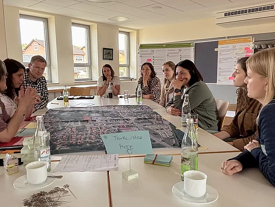Mehrere Personen sitzen an einem Tisch um einen großen Luftbild-Stadtplan herum und diskutieren; Notizen, Getränke und Arbeitsmaterialien liegen auf dem Tisch.