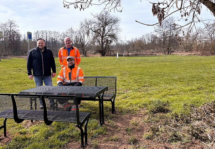 Drei Männer, zwei davon in orangefarbener Arbeitskleidung, stehen und sitzen an einer neuen Sitzgruppe mit Tisch auf einer Wiese unter einem Baum, während im Hintergrund eine offene Landschaft mit weiteren Bäumen zu sehen ist.