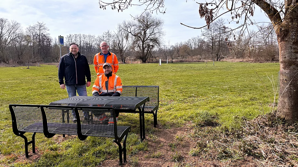 Drei Männer, zwei davon in orangefarbener Arbeitskleidung, stehen und sitzen an einer neuen Sitzgruppe mit Tisch auf einer Wiese unter einem Baum, während im Hintergrund eine offene Landschaft mit weiteren Bäumen zu sehen ist.
