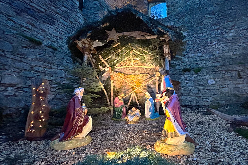 Das Bild zeigt eine stimmungsvoll beleuchtete Weihnachtskrippe mit lebensgroßen Figuren der Heiligen Familie und der Heiligen Drei Könige, aufgestellt in einer steinernen Ruine unter einem mit Tannenzweigen geschmückten Stall.