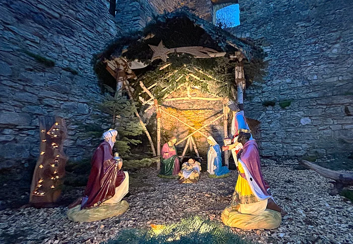 Das Bild zeigt eine stimmungsvoll beleuchtete Weihnachtskrippe mit lebensgroßen Figuren der Heiligen Familie und der Heiligen Drei Könige, aufgestellt in einer steinernen Ruine unter einem mit Tannenzweigen geschmückten Stall.