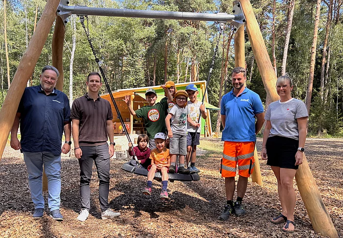 Mehrere Kinder sitzen und stehen auf einer neuen Nestschaukel auf dem Gelände des BRK-Waldkindergartens „Zwergenwald“ in Teublitz. Daneben stehen Bürgermeister Thomas Beer, der städtische Sachbearbeiter für Kinderbetreuung Thomas Fyrguth, Bauhofleiter Peter Roidl und Kindergartenleiterin Damaris Heymann. Im Hintergrund sind die Schutzhütte des Waldkindergartens sowie der umliegende Wald zu sehen.