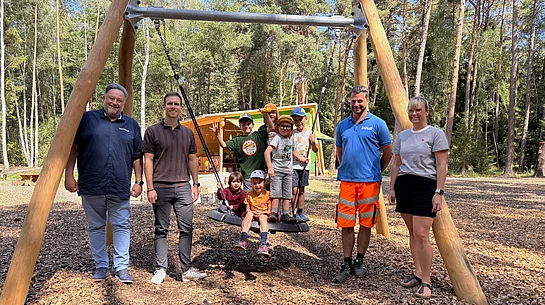 Mehrere Kinder sitzen und stehen auf einer neuen Nestschaukel auf dem Gelände des BRK-Waldkindergartens „Zwergenwald“ in Teublitz. Daneben stehen Bürgermeister Thomas Beer, der städtische Sachbearbeiter für Kinderbetreuung Thomas Fyrguth, Bauhofleiter Peter Roidl und Kindergartenleiterin Damaris Heymann. Im Hintergrund sind die Schutzhütte des Waldkindergartens sowie der umliegende Wald zu sehen.