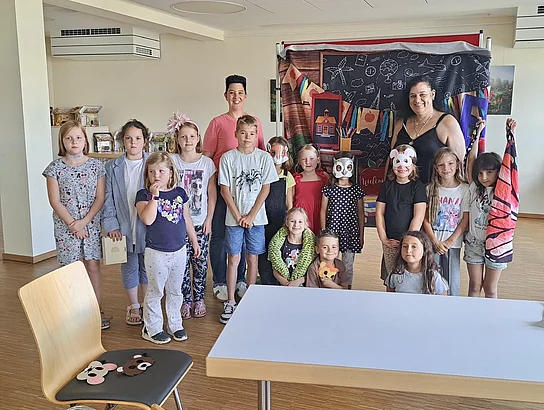 Gruppenfoto der Theateraktion im Sommerferienprogramm 2026. Mehrere Kinder im Grundschulalter stehen gemeinsam mit zwei erwachsenen Betreuerinnen in einem Raum des Mehrgenerationenhauses Saltendorf. Einige Kinder tragen selbst gestaltete Tiermasken oder halten Requisiten in den Händen. Hinter der Gruppe steht eine farbenfrohe Theaterkulisse mit Schultafel, Büchern und weiteren Motiven. Die Kinder nahmen an der Aktion „Komm, lass uns Theater spielen“ mit dem Partykistl teil.