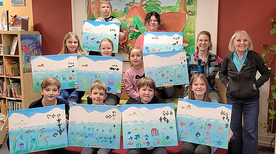 Eine Gruppe von Kindern präsentiert gemeinsam mit zwei Erwachsenen in einer Bücherei stolz ihre selbst gestalteten Bilder mit bunten Unterwasserlandschaften, auf denen Fische, Quallen und andere Meeresmotive zu sehen sind.