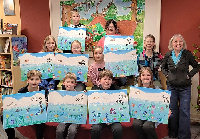 Eine Gruppe von Kindern präsentiert gemeinsam mit zwei Erwachsenen in einer Bücherei stolz ihre selbst gestalteten Bilder mit bunten Unterwasserlandschaften, auf denen Fische, Quallen und andere Meeresmotive zu sehen sind.
