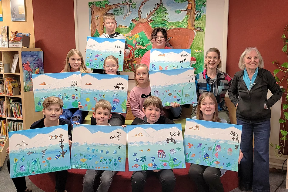Eine Gruppe von Kindern präsentiert gemeinsam mit zwei Erwachsenen in einer Bücherei stolz ihre selbst gestalteten Bilder mit bunten Unterwasserlandschaften, auf denen Fische, Quallen und andere Meeresmotive zu sehen sind.
