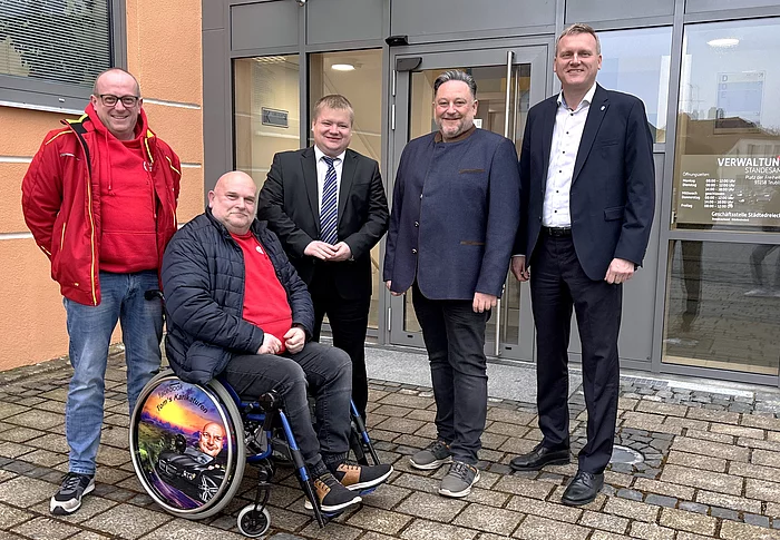 Vor dem Eingang eines Verwaltungsgebäudes stehen mehrere Männer zusammen, darunter eine Person im Rollstuhl, und posieren lächelnd für ein gemeinsames Gruppenfoto.