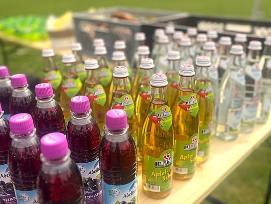 Auf einem Tisch stehen verschiedene Getränke in Flaschen, darunter Apfelschorle und Johannisbeergetränke, bereit für die Gäste der Veranstaltung.