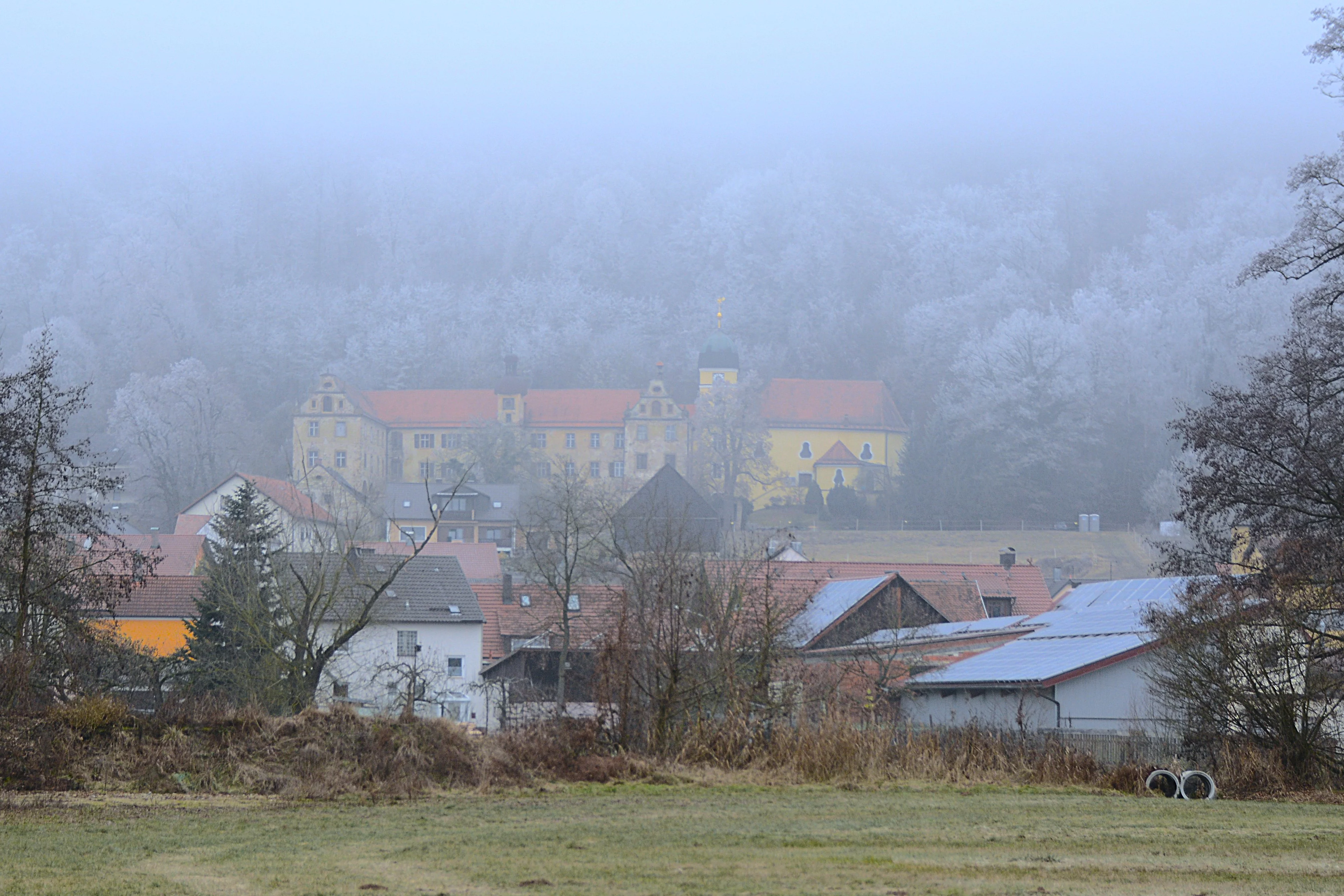 Eine prächtige Schlossanlage thront über dem Dorf an einem winterlichen nebelverhangenen Morgen.